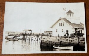 SKU-3231::  Postcard RPPC: amphibious aircraft, Model-T, American Flag, New England Waterfront. Unposted. Fine condition. Real Photo.